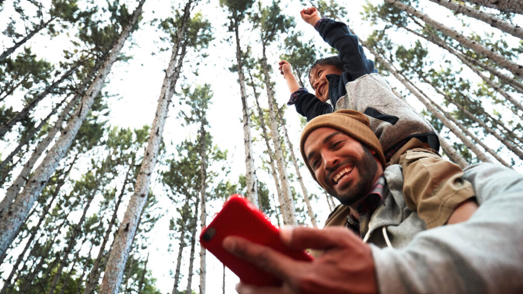 Father smiling and giving child shoulder ride in the forest while looking at smartphone in hand