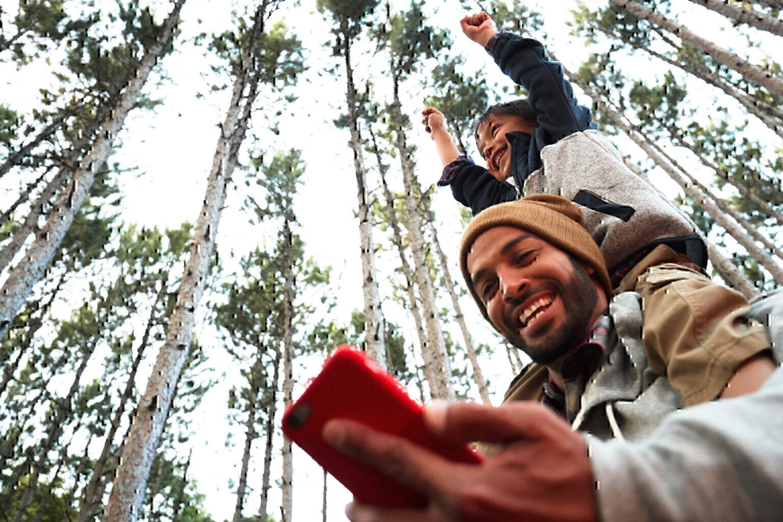 Father smiling and giving child shoulder ride in the forest while looking at smartphone in hand