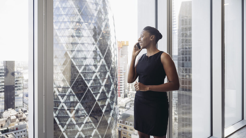 Smiling woman standing at the window of a high-rise office and talking on the phone