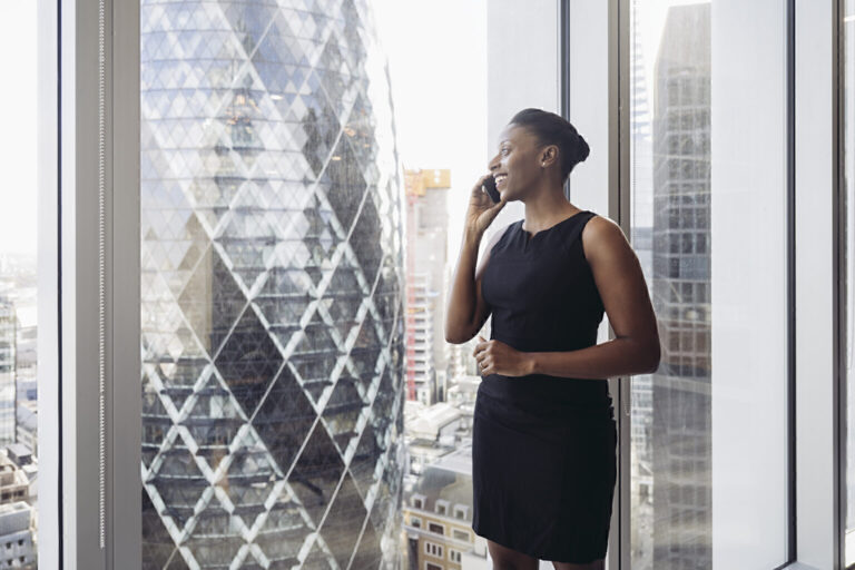 Smiling woman standing at the window of a high-rise office and talking on the phone