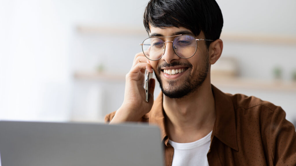 A young man in glasses smiles while looking at his laptop and talking on the phone.