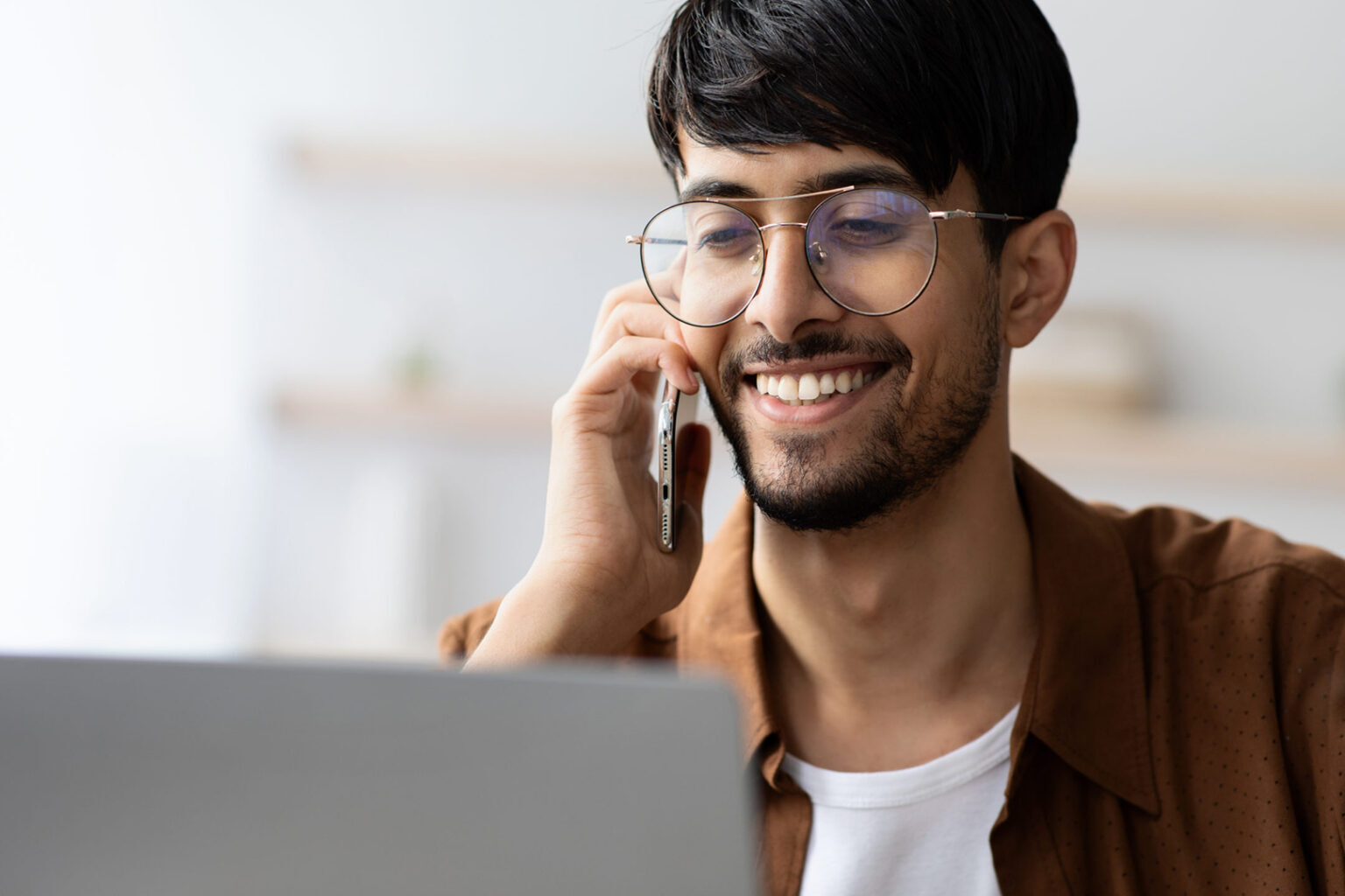 A young man in glasses smiles while looking at his laptop and talking on the phone.