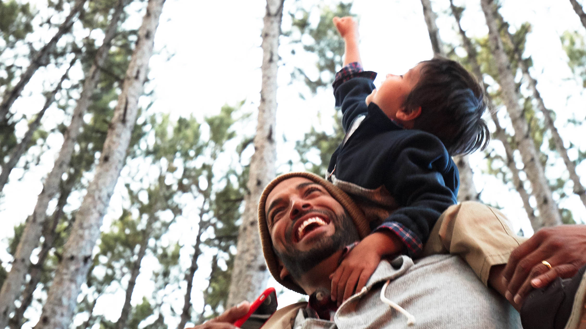 Father smiling and giving child shoulder ride in the forest while holding smartphone in hand