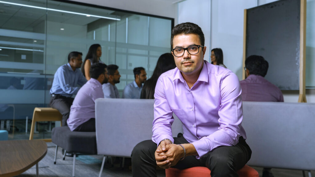 A male CSG employee sits in the Bangalore office as colleagues gather for discussion in the background.