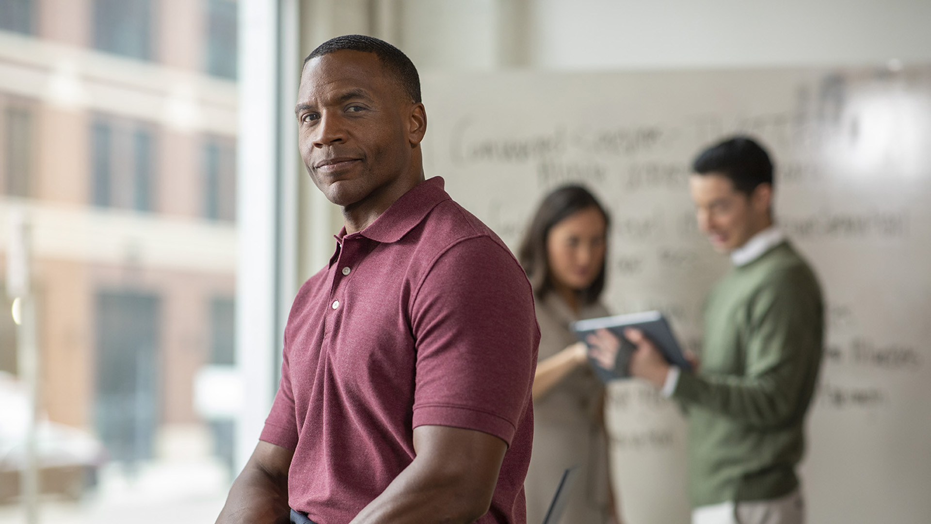 An African-American businessman wearing a red shirt smiles at the camera while two colleagues talk in the background.