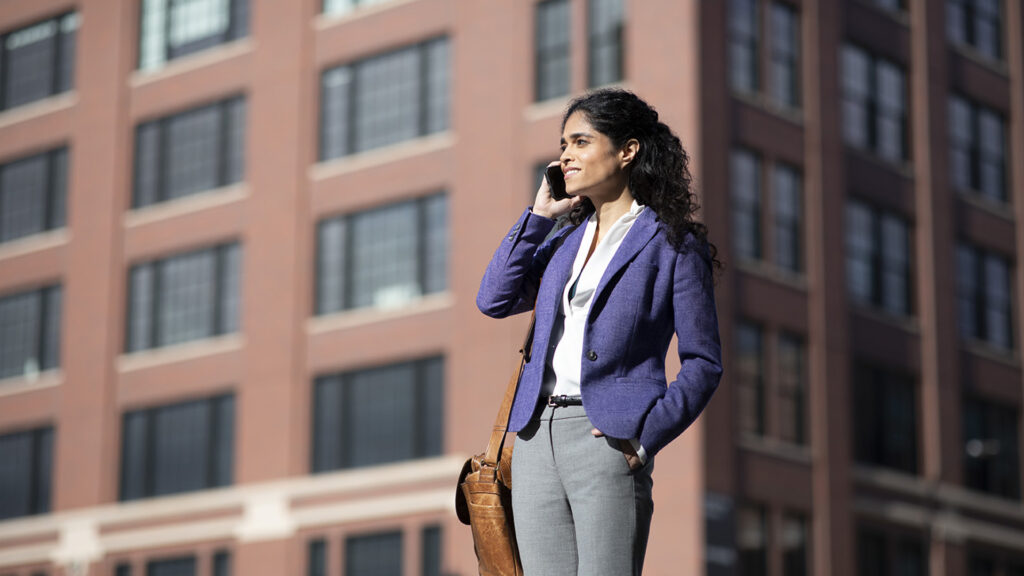 A businesswoman in a purple jacket talks on her phone while standing in front of a brown building.