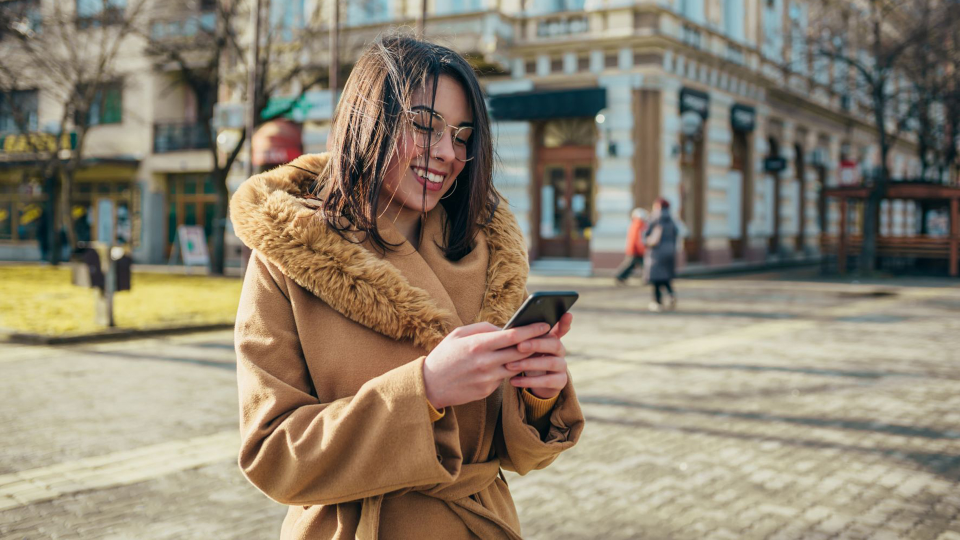 A young woman wearing a brown winter jacket reacts happily to a message she received on her phone.