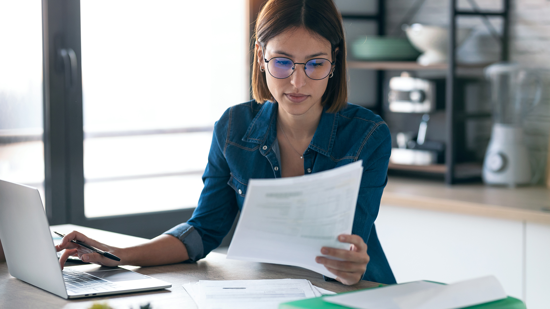 Woman working on her computer while consulting some invoices and documents in her kitchen at home.