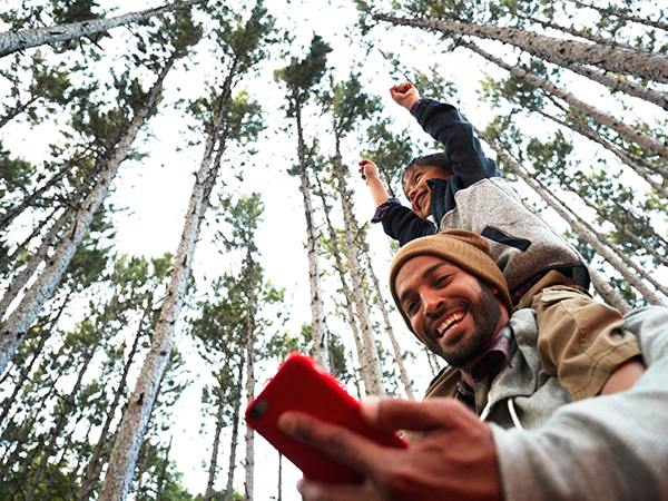 A dad giving his son a piggyback ride on a hike smiles down at his phone