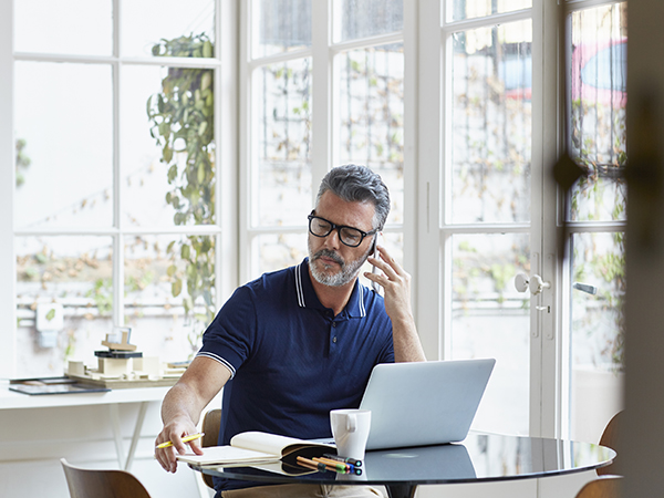 Mature businessman using mobile phone while writing notes at table in office