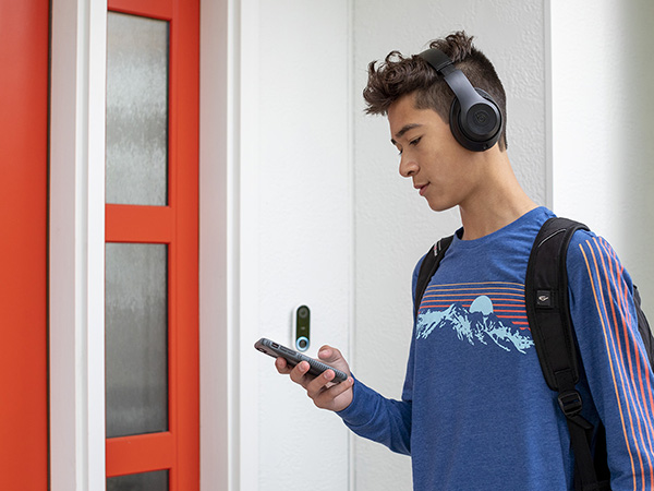 A teenage boy wearing a blue shirt, backpack, and headphones checks his phone while entering his house