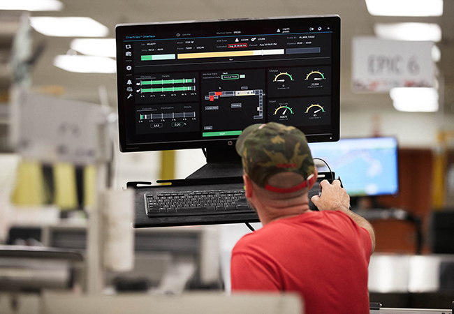 Man wearing a baseball hat is facing away from the camera and looking at a monitor in a distribution center.
