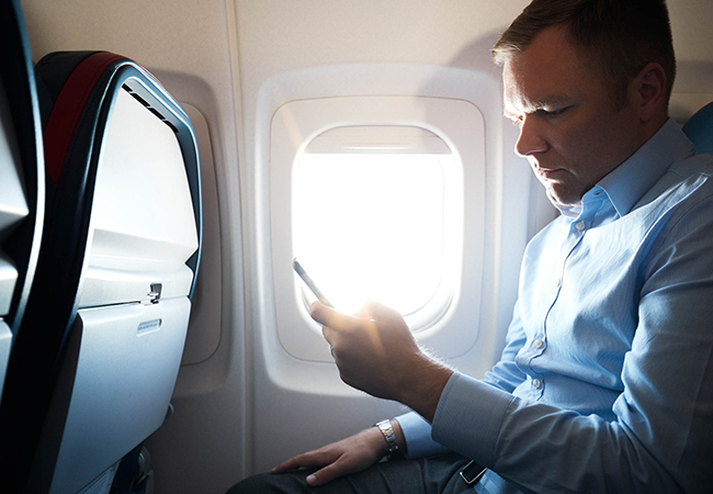 A man sitting in a window seat on an airplane looks at his phone.