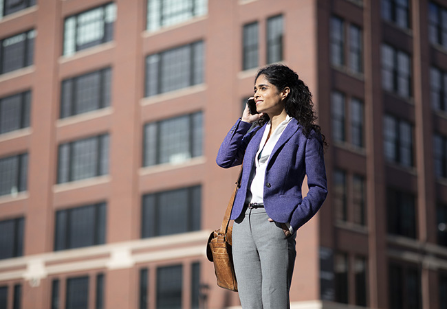 A businesswoman in a purple jacket talks on her phone while standing in front of a brown building.