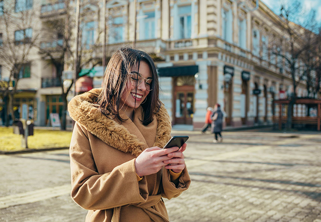 A young woman wearing a brown winter jacket reacts happily to a message she received on her phone.