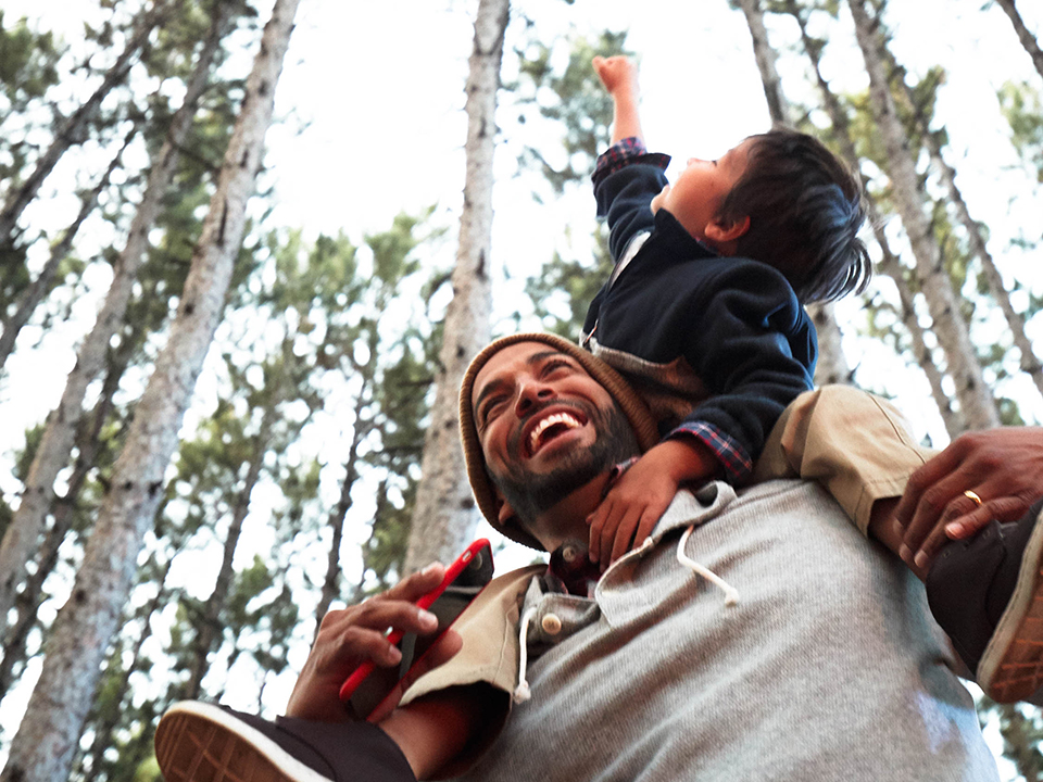 Father smiling and giving child shoulder ride in the forest while holding smartphone in hand