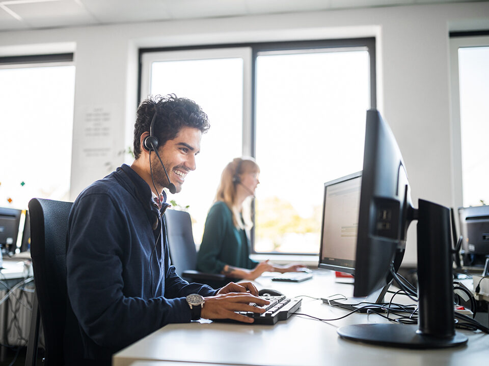 A smiling customer service representative is using a computer at his desk