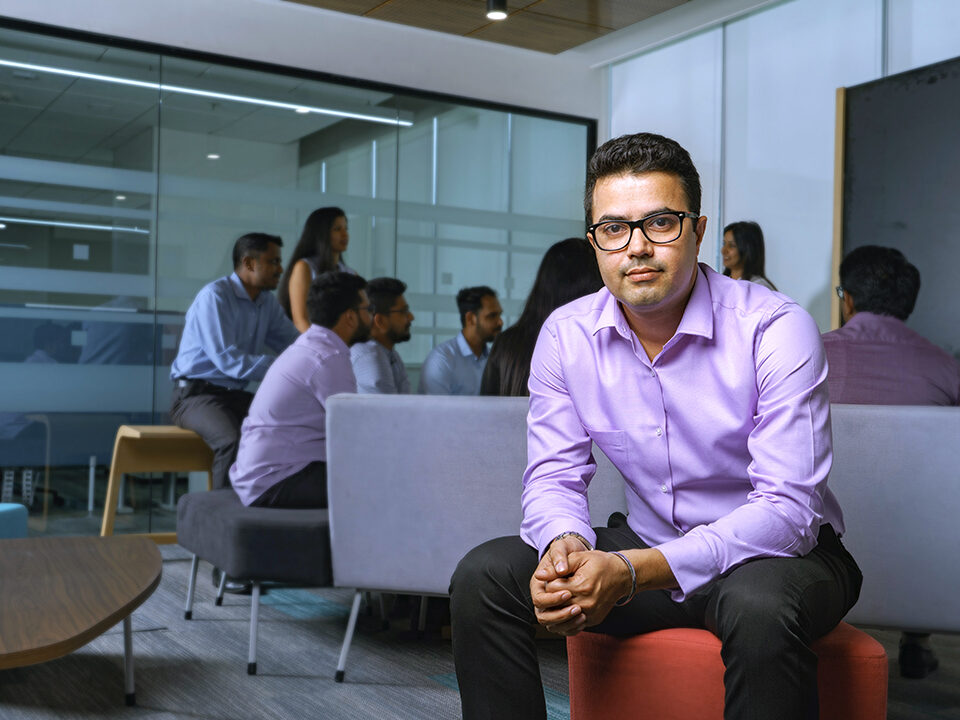 A male CSG employee sits in the Bangalore office as colleagues gather for discussion in the background.