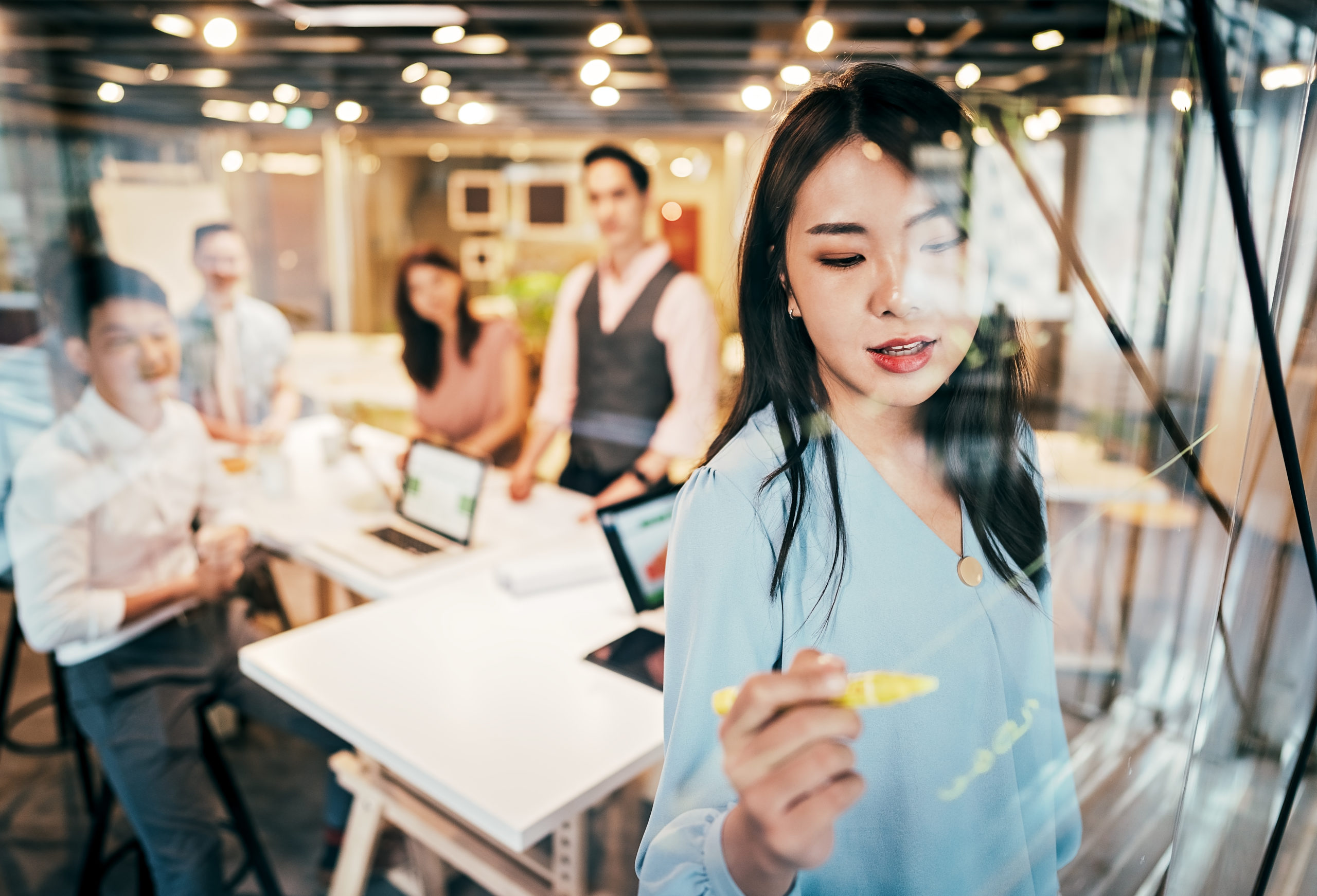 Young Group Of Professionals Developing Business Strategy and Providing The Business Plan. In front stands a woman writing on a clear draw board with a yellow marker.