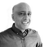 Professional headshot of a bald man with a warm smile, wearing a dark collared shirt.