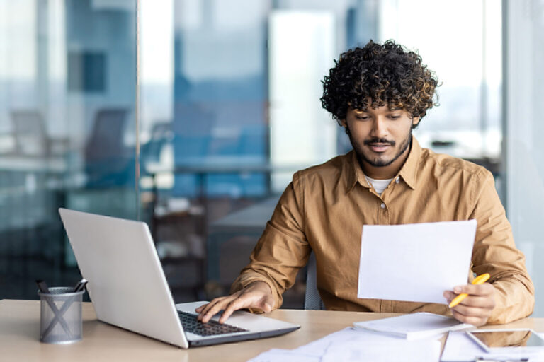 A young businessman with curly hair sits in an office, immersed in his laptop, crafting a digital