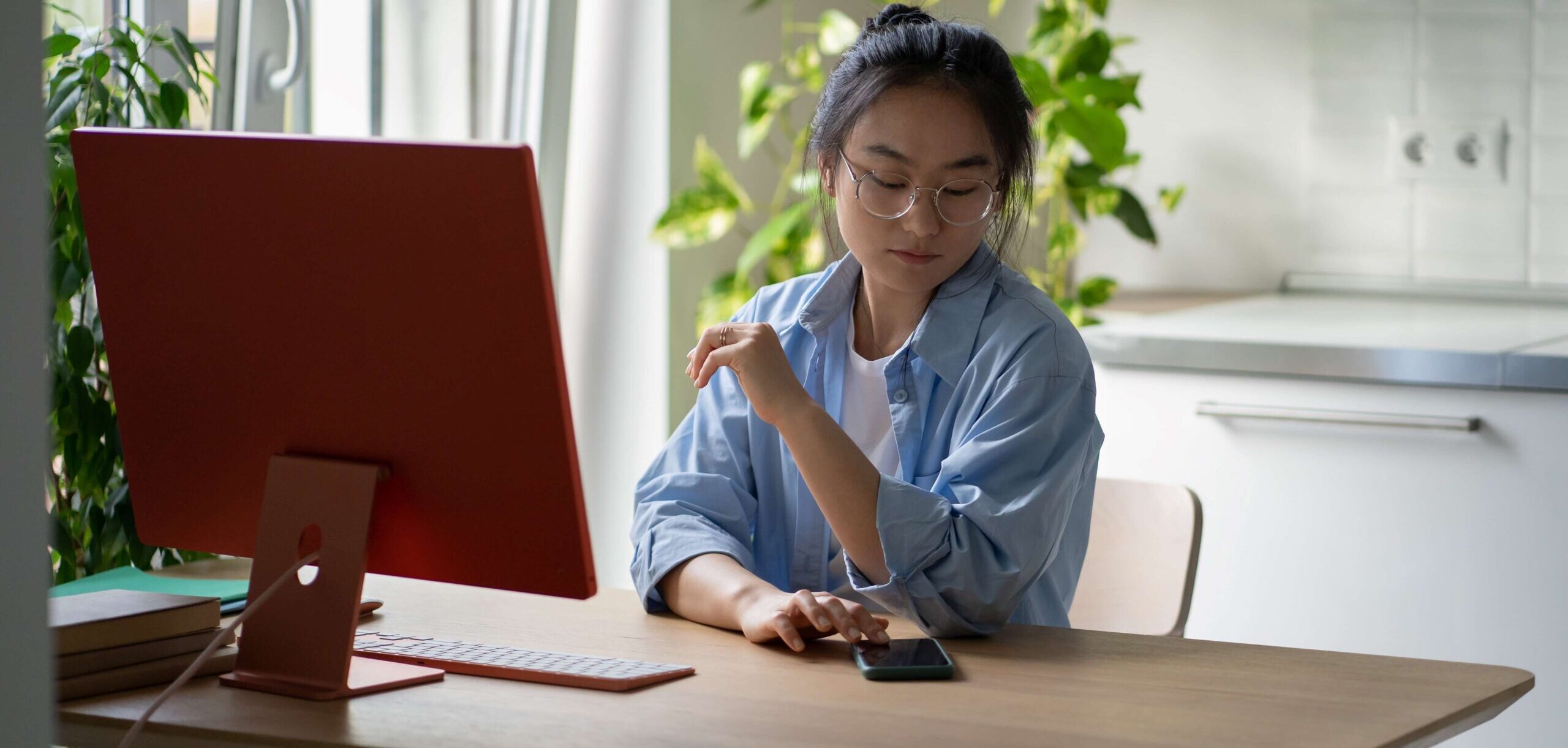 A student checks her phone while studying on her desktop computer at home