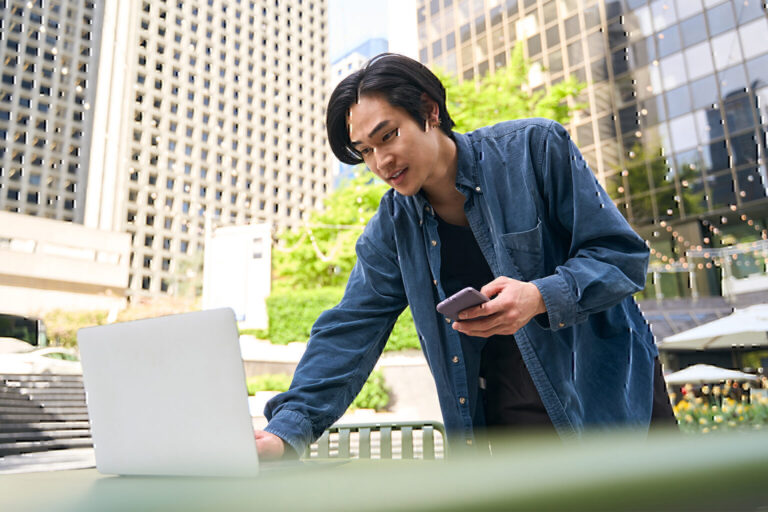 Brunette asian man checking laptop email and holding mobile phone