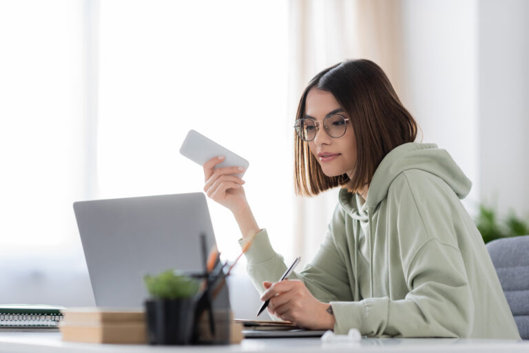 Brunette freelancer in eyeglasses holding cellphone and writing on notebook near laptop at home