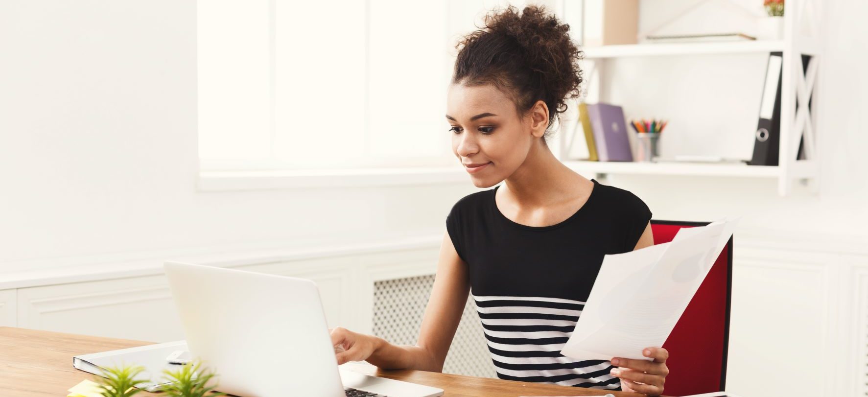 A woman with an eager expression inputs commands on her laptop while holding a paper document in one hand