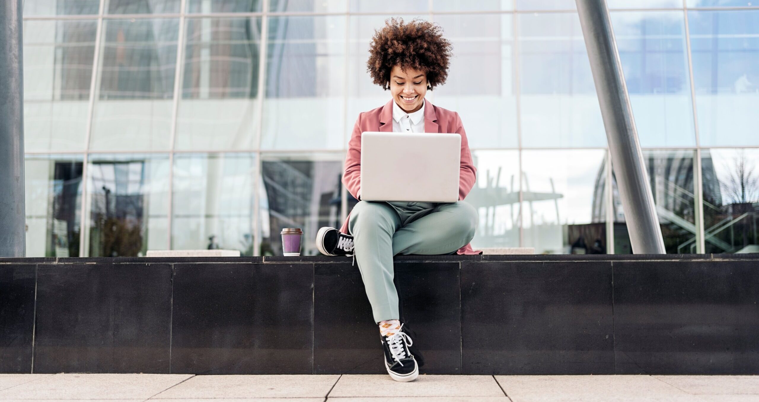 A woman in a pink blazer and jeans works on her laptop while sitting in a common area outside an office building.