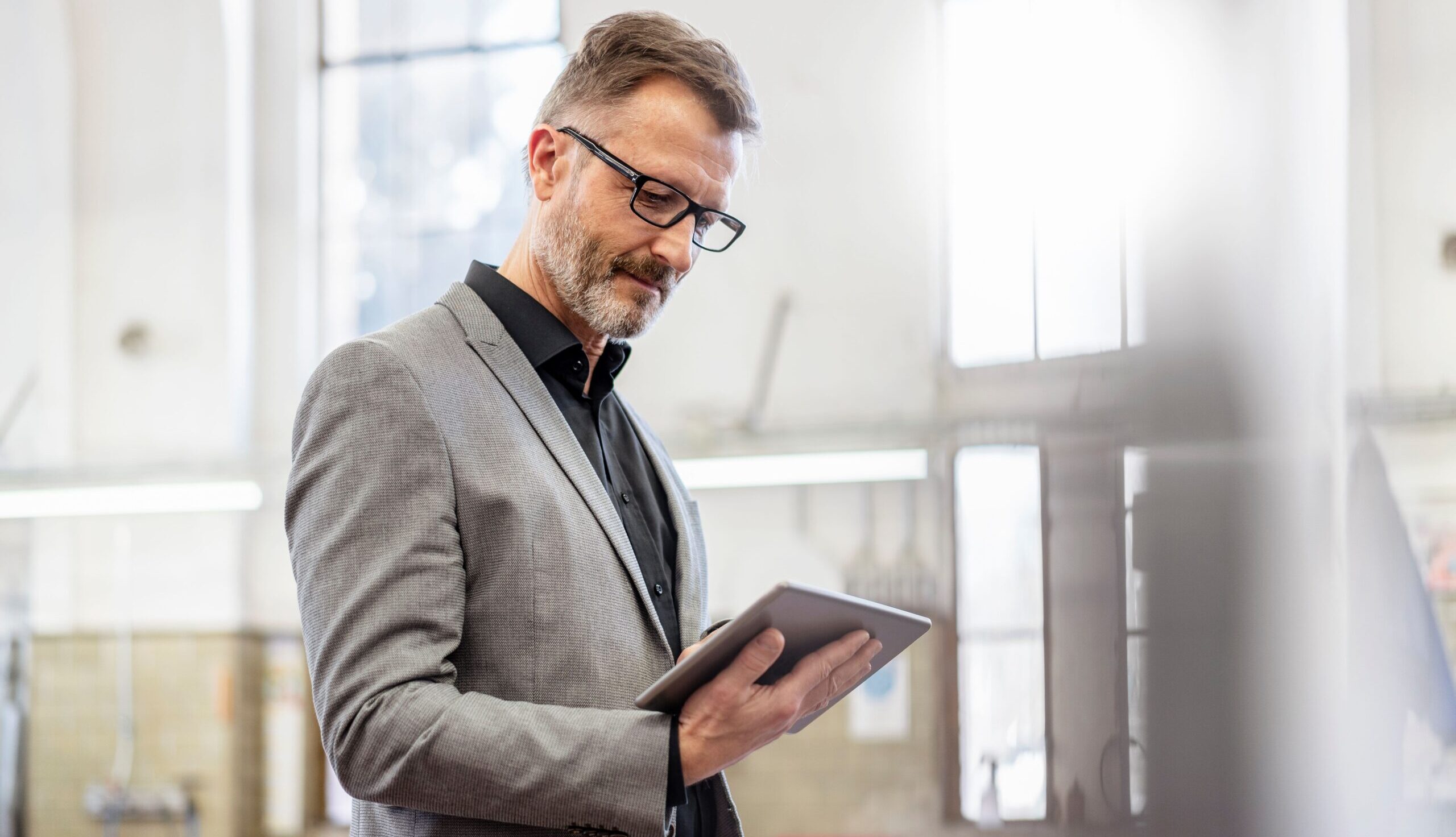 A glasses-wearing businessman reads a tablet while standing on a factory floor.