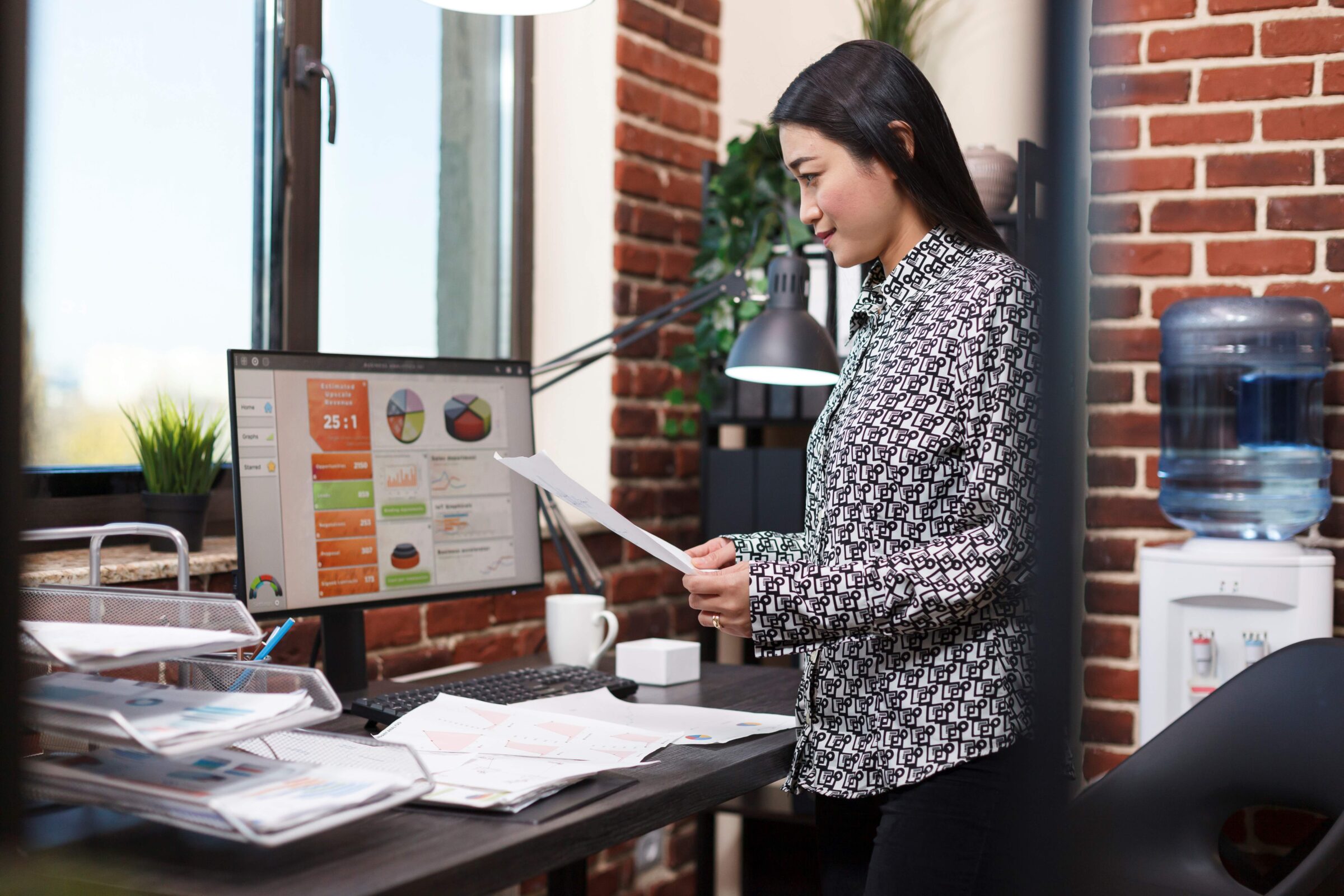 A female professional reviews business documents while in front of her desktop computer screen, which shows an analytics dashboard.