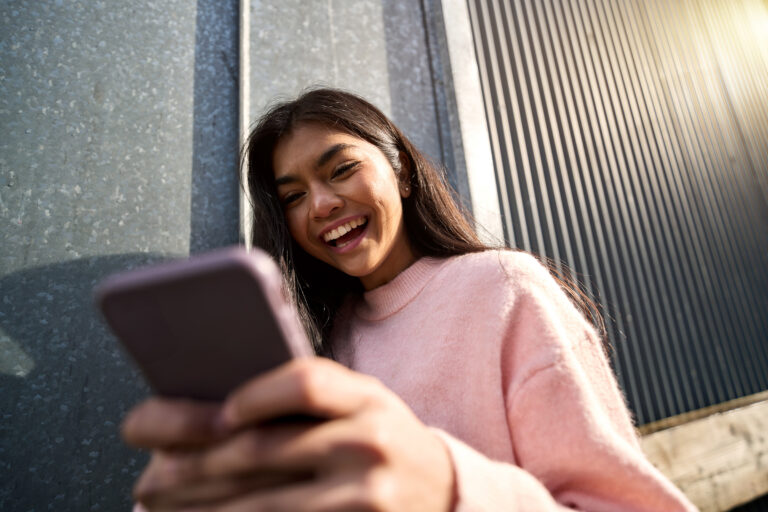 Happy cheerful Asian teen girl blogger using mobile phone standing on street.