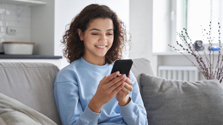 Happy hispanic teen girl holding cell phone using smartphone at home