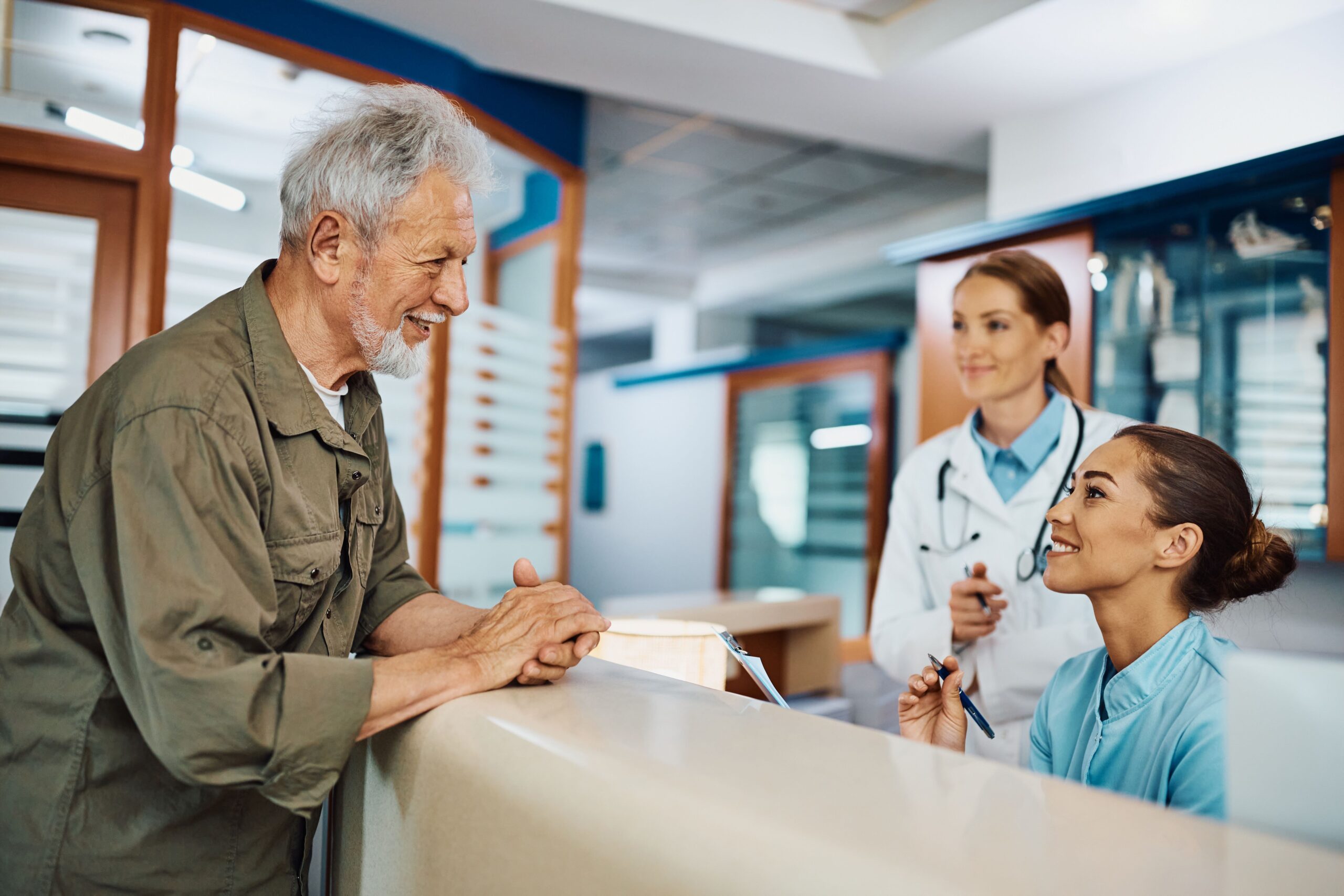 A pleased senior man at a healthcare clinic talks with a receptionist as a smiling female doctor looks on