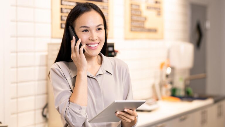 Young cheerful waitress or manager of restaurant communicating with client on mobile phone while using digital tablet by workplace