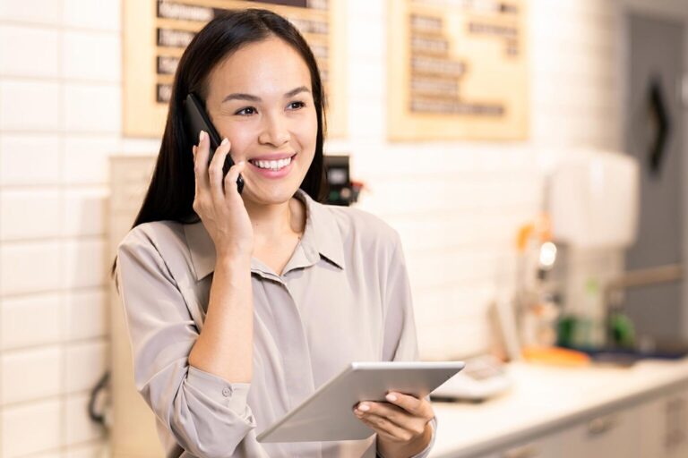 Young cheerful waitress or manager of restaurant communicating with client on mobile phone while using digital tablet by workplace