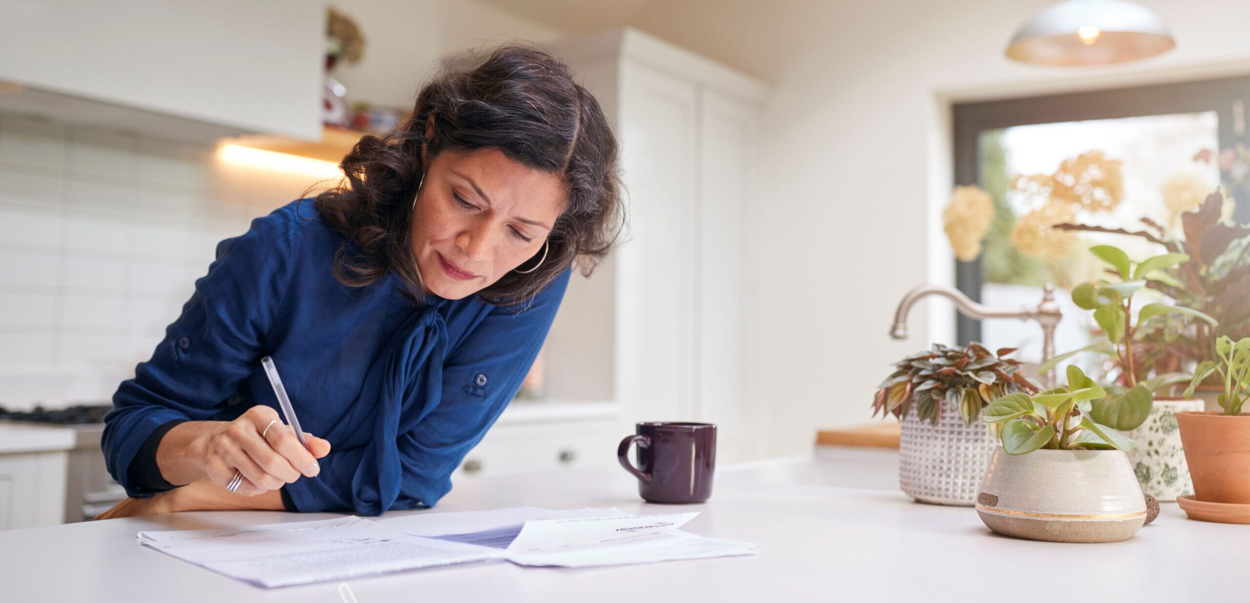 A woman leans over her kitchen counter, holding a pen and reviewing a set of paper documents.