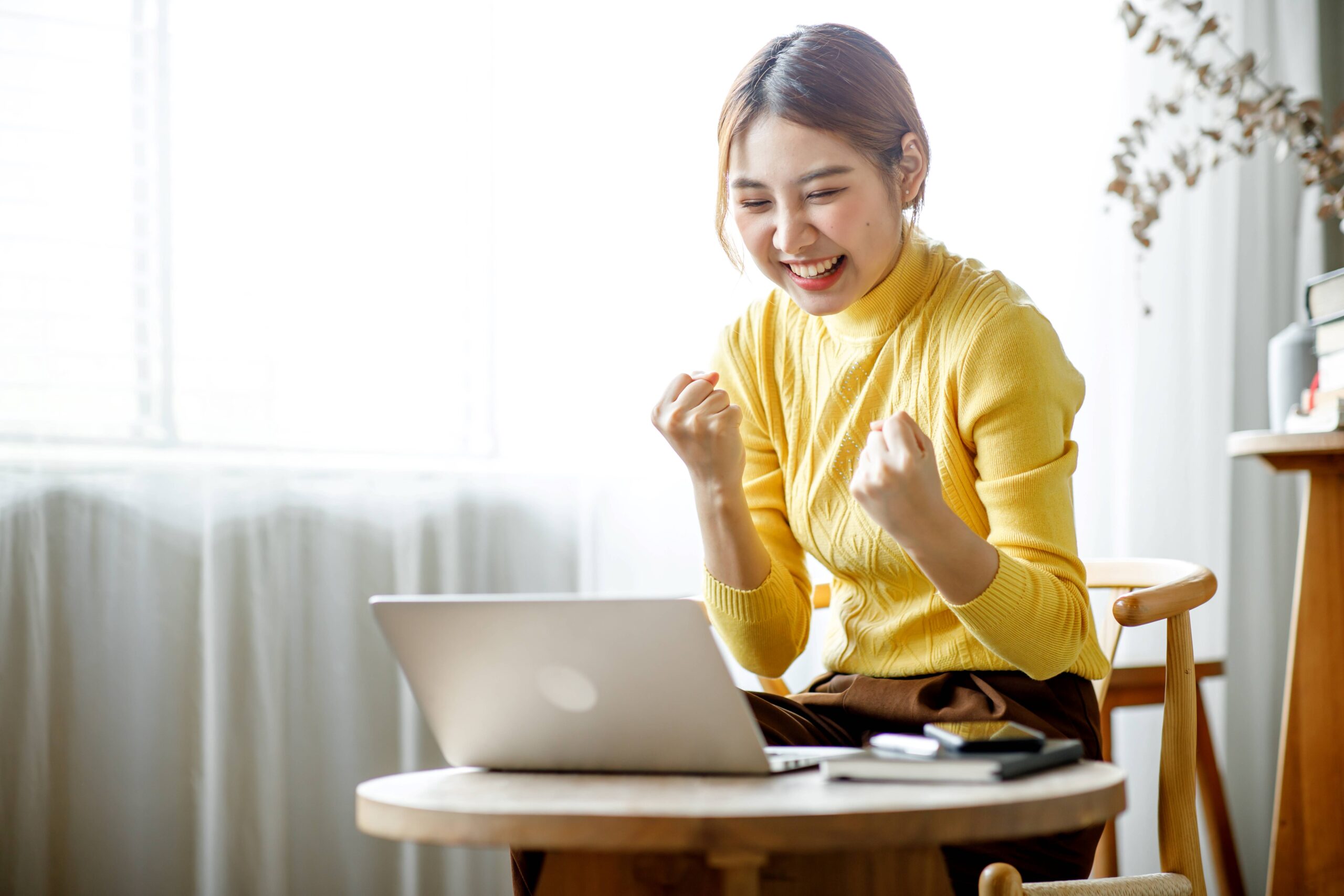 A smiling young woman clenches her fists in triumph while looking at her laptop.