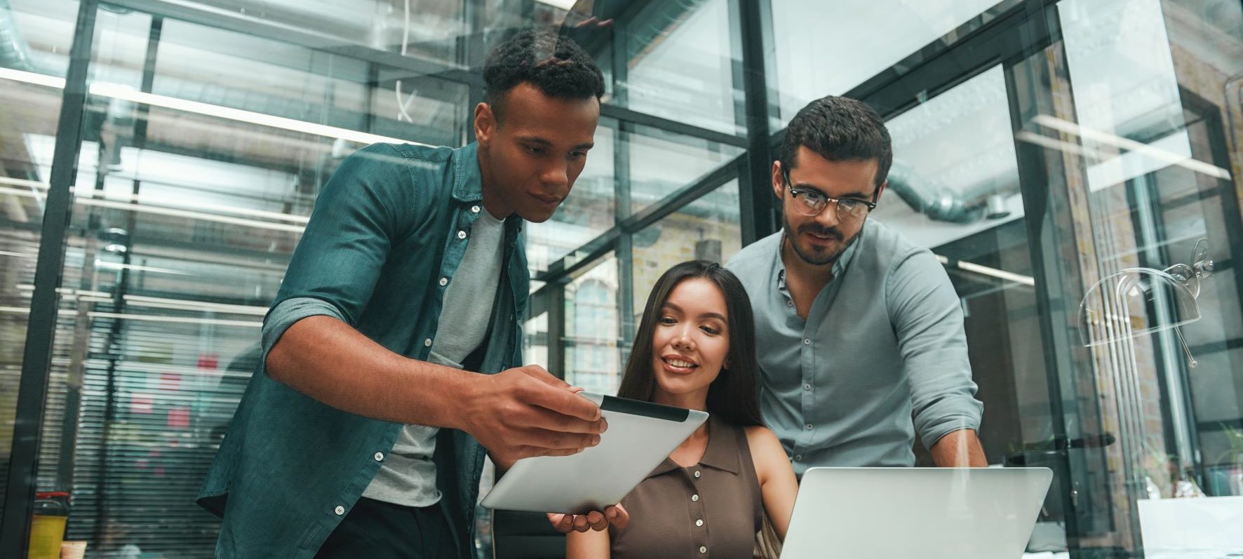 A team of three young professionals collaborating on a project using a tablet and desktop computer.