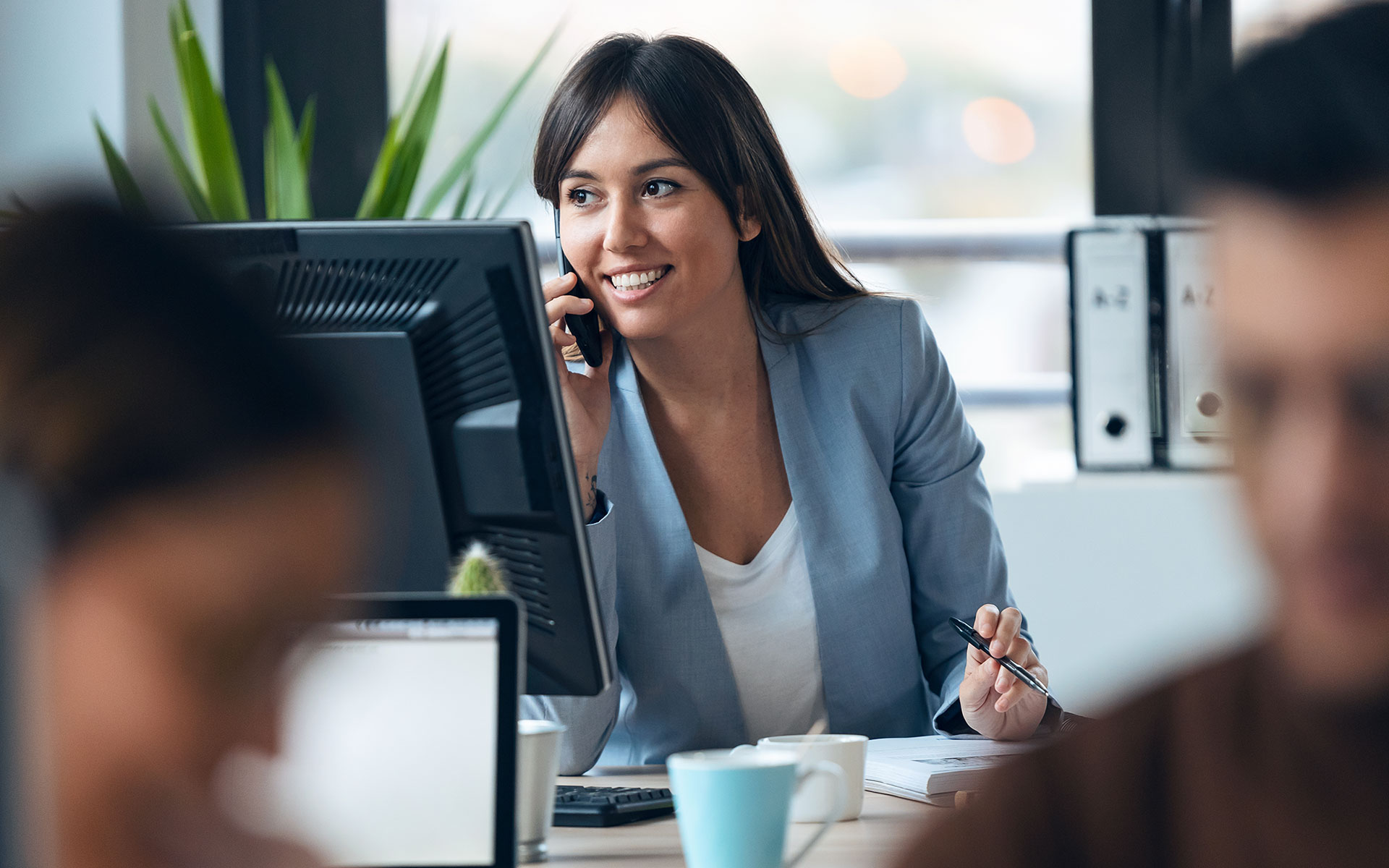 Smart young businesswoman talking with mobile phone while working with computer in modern workspace