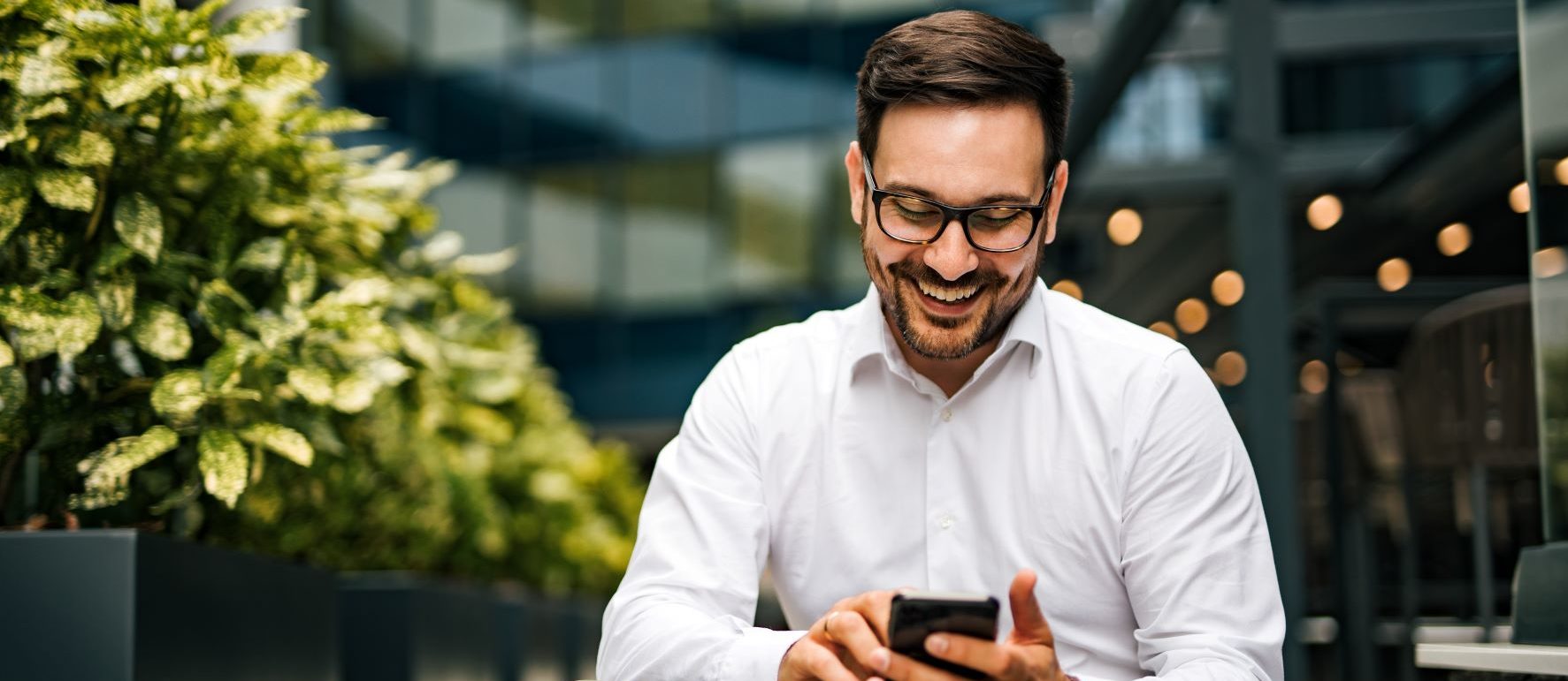 Man in glasses laughing and looking at his smartphone outdoors.