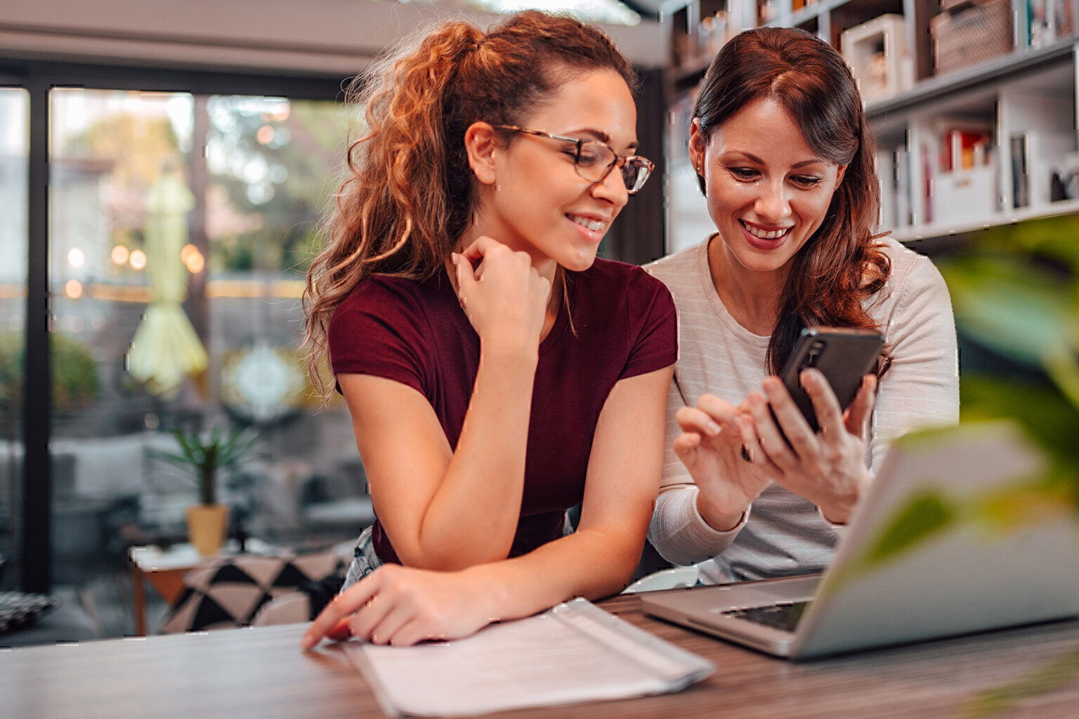 Two women share phone and laptop in coffee shop