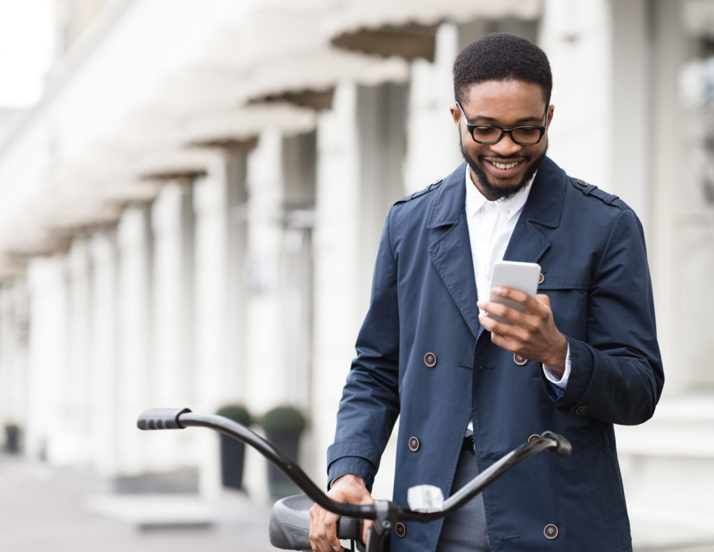 Young Black man texting on phone and standing with bike on city street.