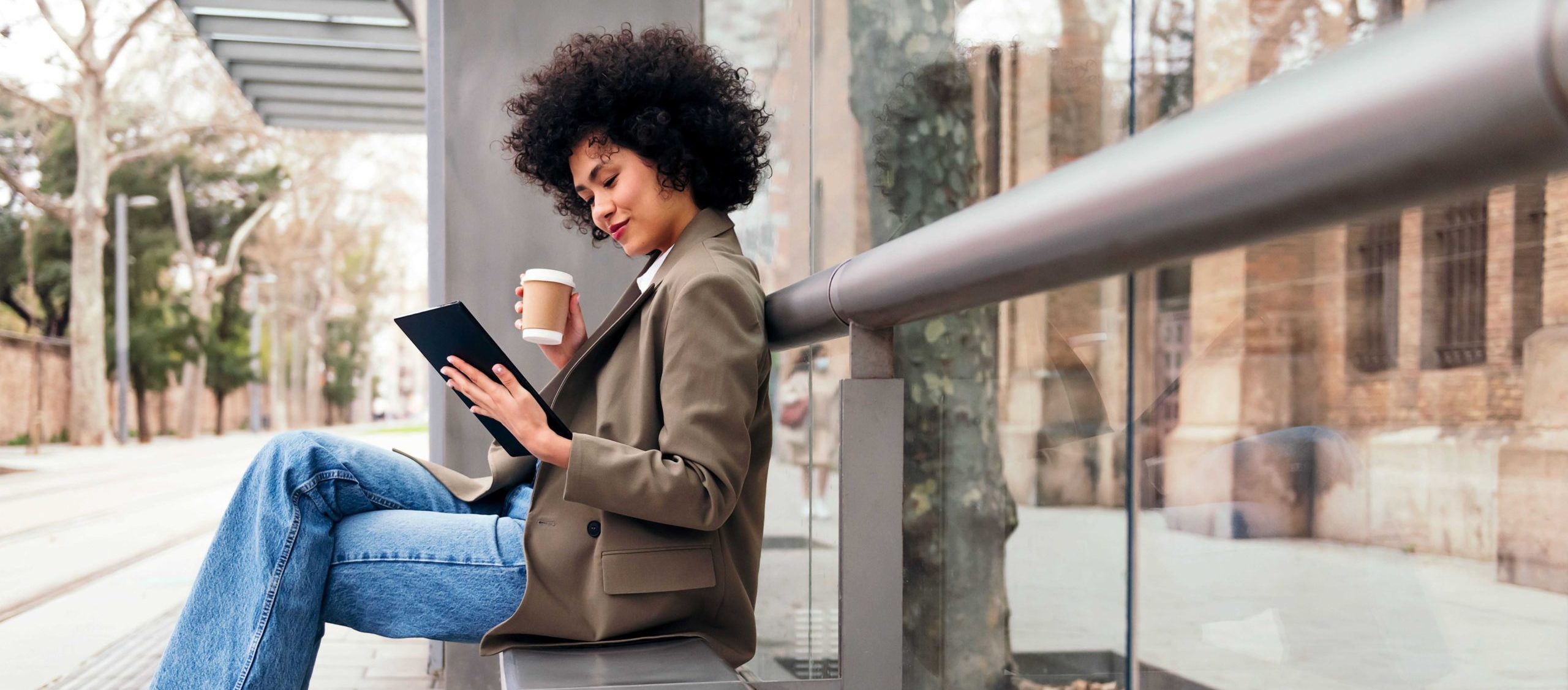 A woman relaxes at a bus stop reading a tablet device and holding a cup of coffee.