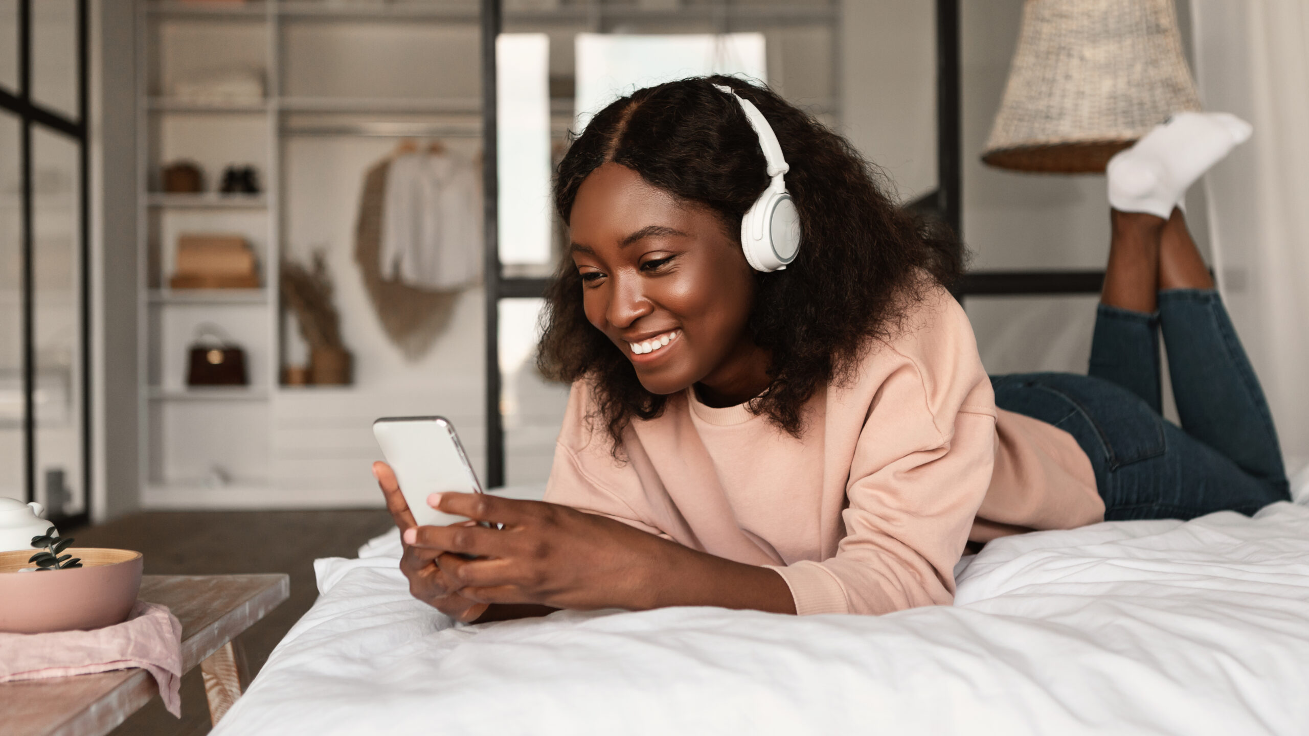 A young African-American girl smiling at her cellphone while wearing headphones and laying on her bed.